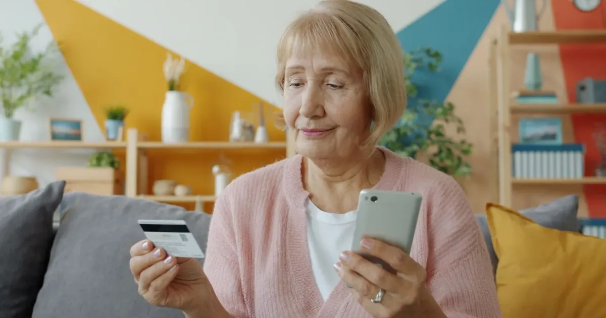 Starší žena kontroluje bankovnictví v telefonu s kartou v ruce